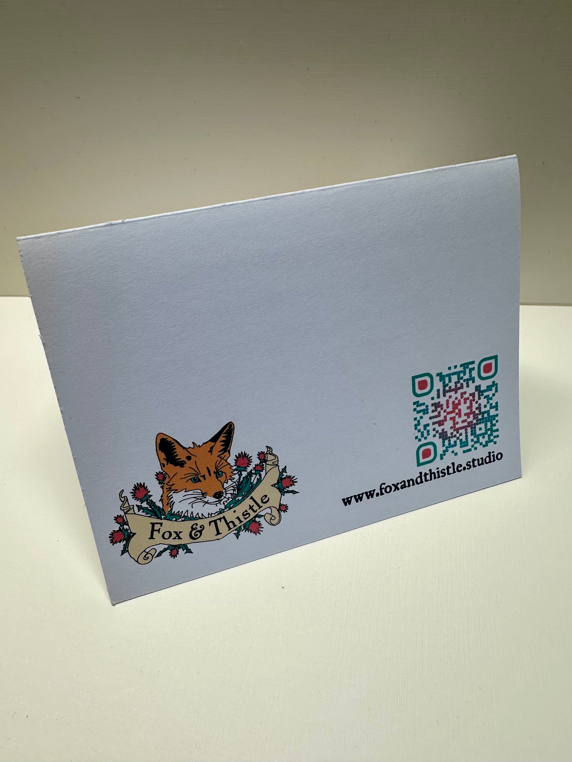 Rear picture of 'Cat's Cradle Card' from Fox & Thistle Studio. Shows the studio's signature Fox surrounded by Thistle, a QR code, and the website address.