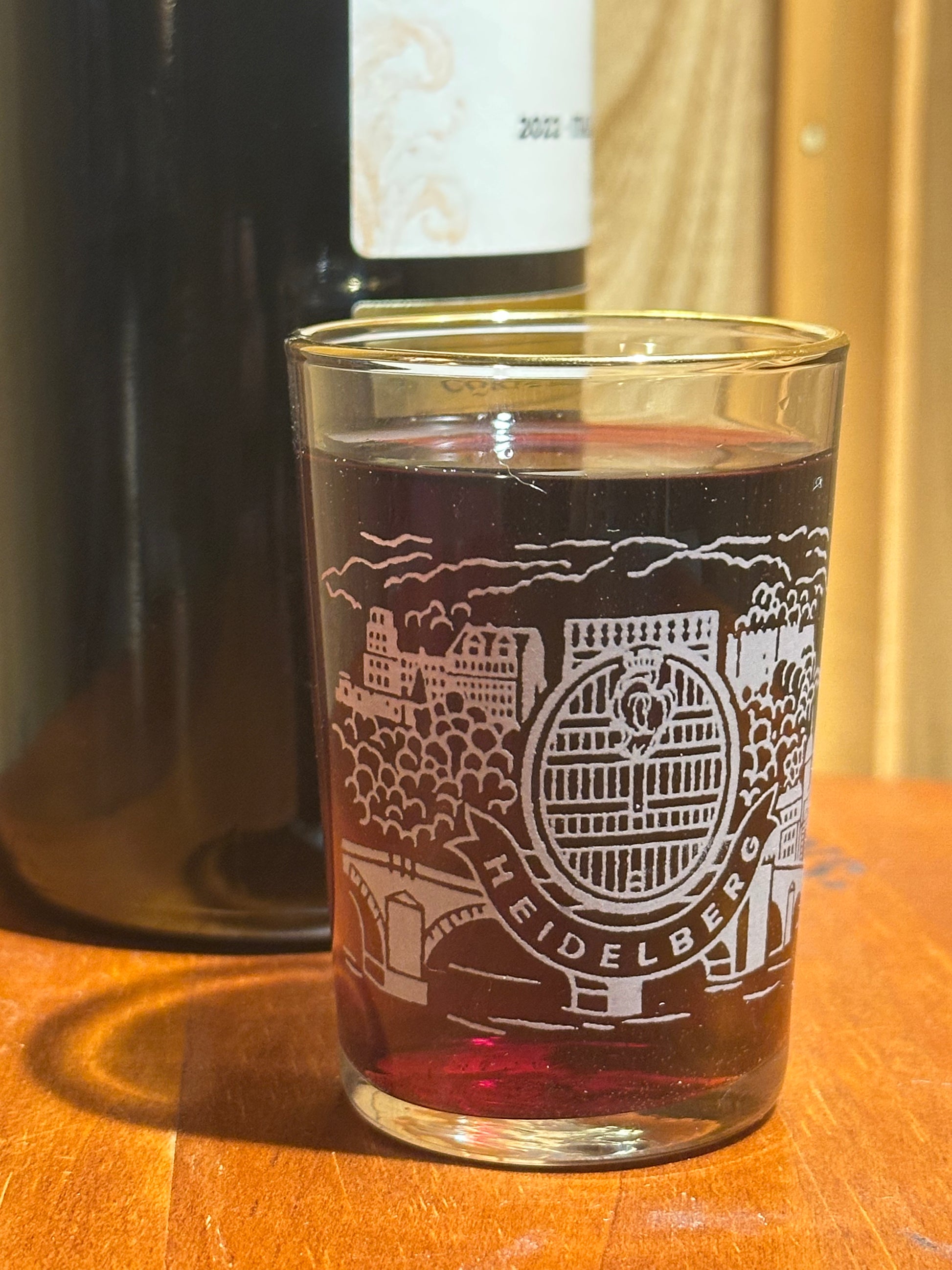 Glass with etched design and red liquid next to a bottle on a wooden surface