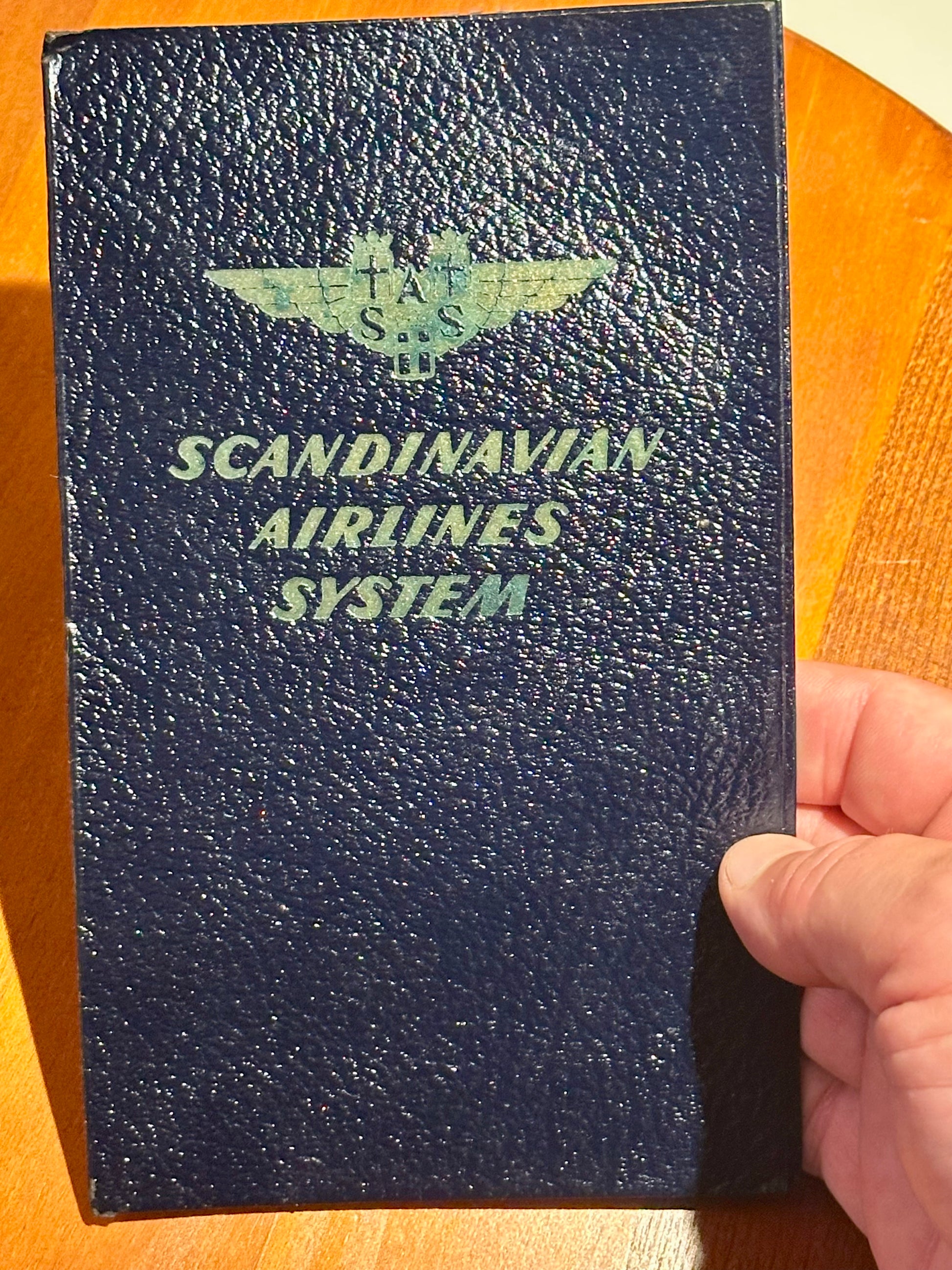 Blue leather-bound book with 'Scandinavian Airlines System' logo held by a hand on a wooden surface.