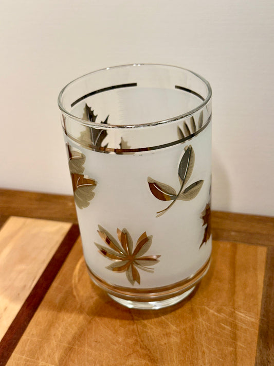 A frosted glass tumbler with a metallic silver botanical leaf pattern, resting on a wooden cutting board against a plain off-white wall.