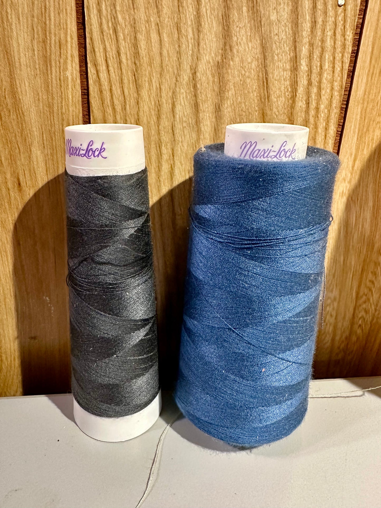Two spools of Maxi-Lock thread, one black and one blue, against a wooden background.