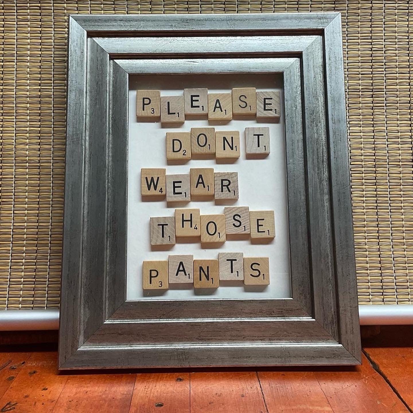 Silver stepped frame with scrabble tiles on a white background spelling out the phrase "Please don't wear those pants."