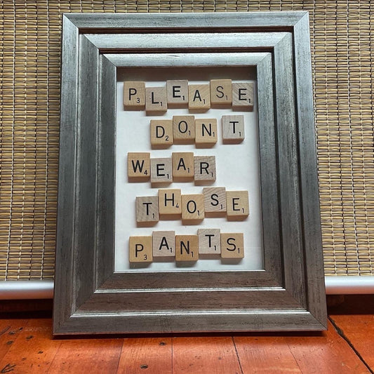 Silver stepped frame with scrabble tiles on a white background spelling out the phrase "Please don't wear those pants."