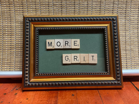 Bronze beaded frame with Scrabble tiles on green background spelling out the phrase "More Grit."