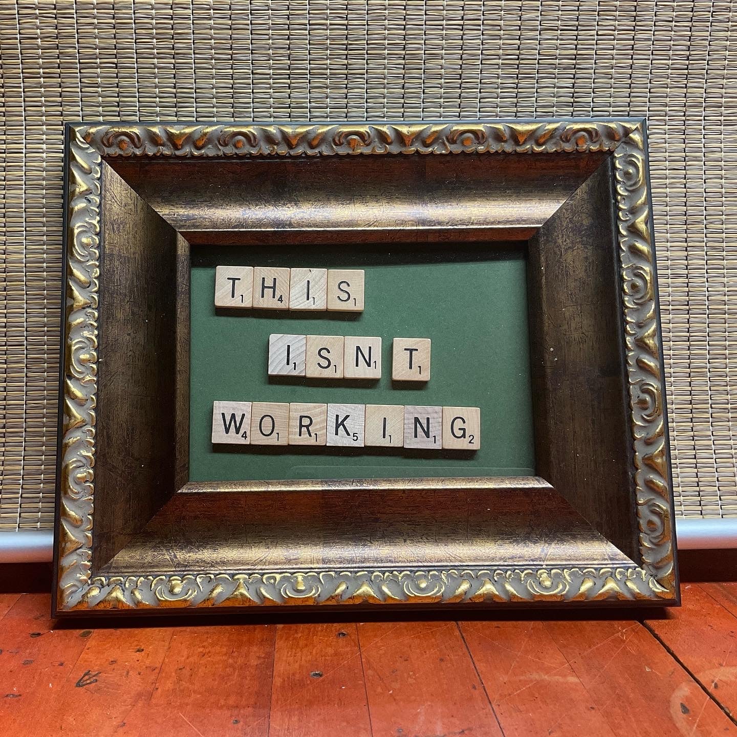 Bronzed frame with scrabble tiles mounted on a green background spelling out the words "this isn't working."