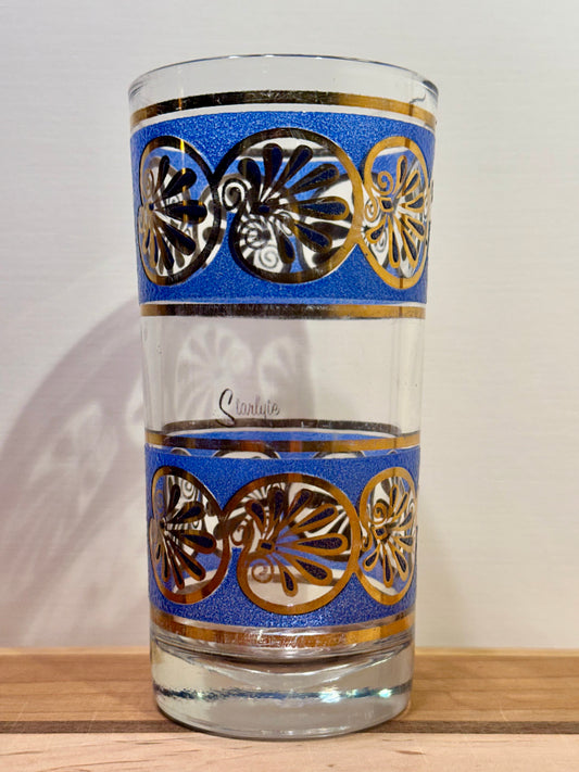 A single clear highball glass tumbler with two decorative bands featuring blue background, gold trim, and seashell motifs, shown on a wooden surface against a light wall.