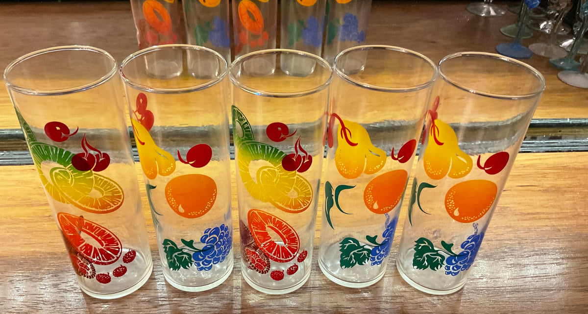 Four clear drinking glasses with colorful fruit prints, including oranges, pears, cherries, and grapes, arranged on a wooden surface.