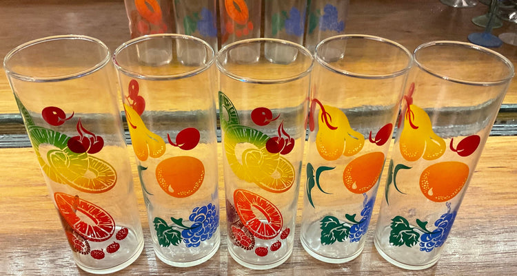 Four clear drinking glasses with colorful fruit prints, including oranges, pears, cherries, and grapes, arranged on a wooden surface.
