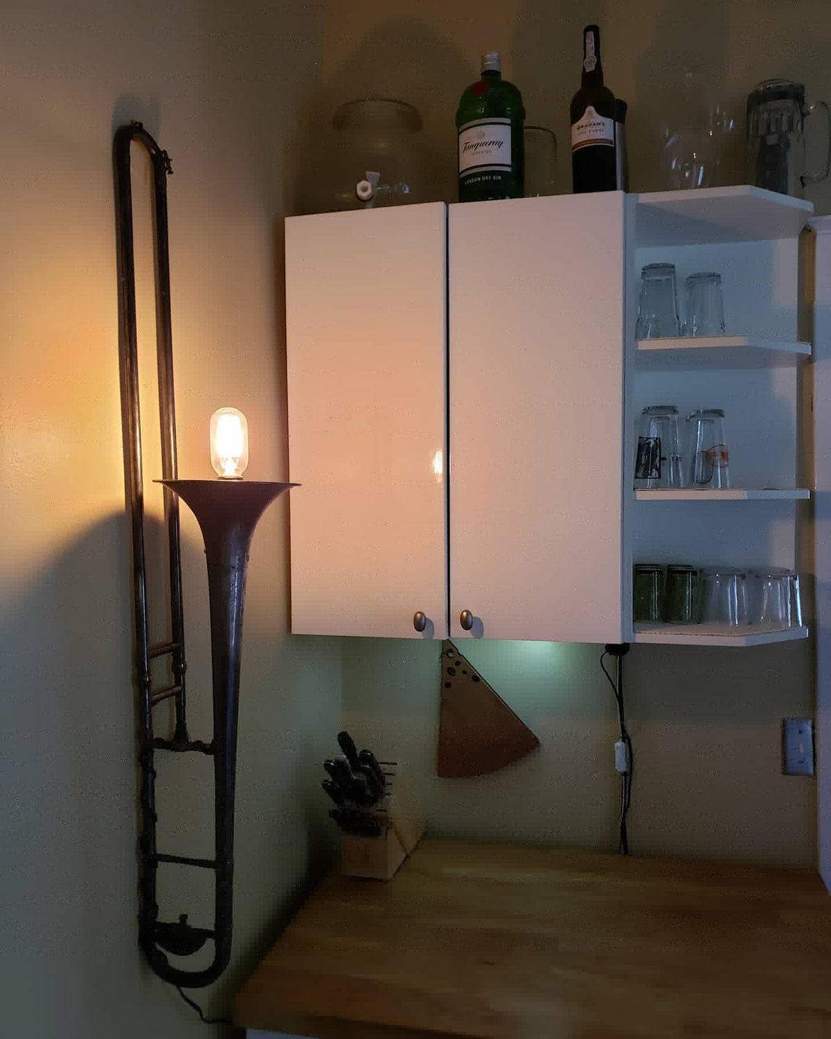 A rustic wall-mounted brass instrument lamp made from a trombone, glowing with a warm Edison bulb, positioned next to a white cabinet with glassware on a wooden counter.