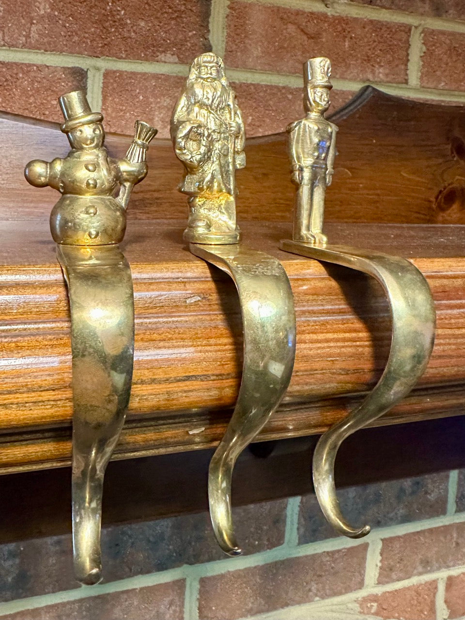 Three decorative brass stocking hooks on mantel with brick background. 