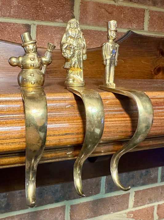 Three decorative brass stocking hooks on mantel with brick background. 