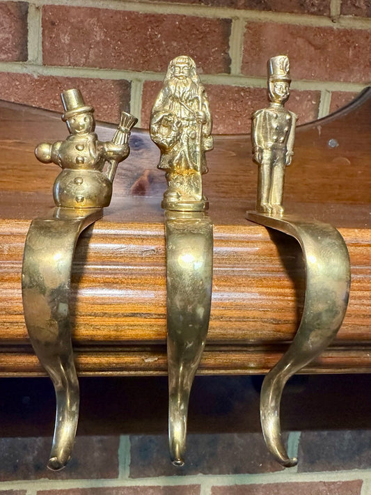 Three decorative brass stocking hooks - snowman, santa, and toy soldier on mantel with brick background.