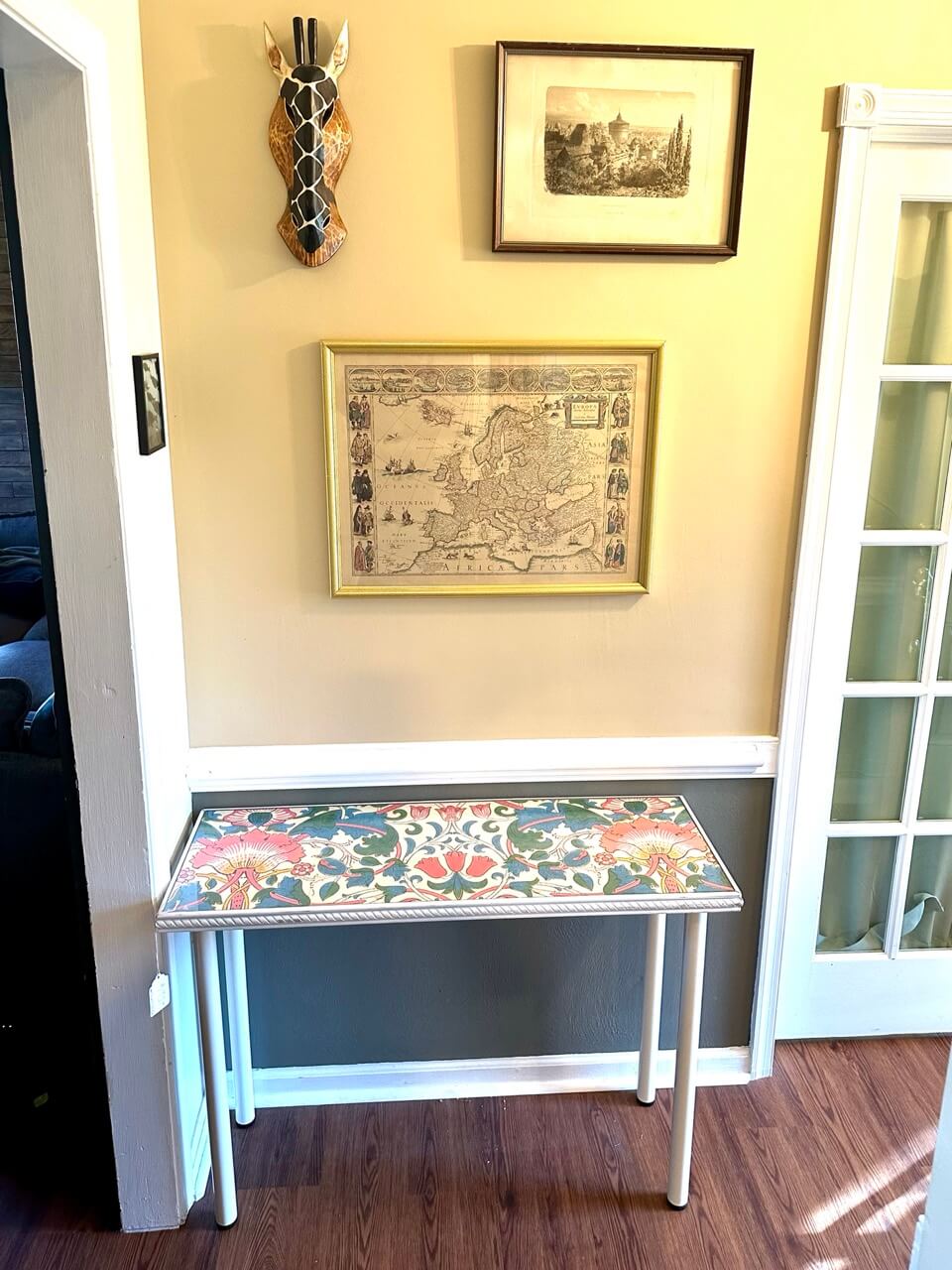 A white floral table in an entry way with a mask and pictures on the wall.