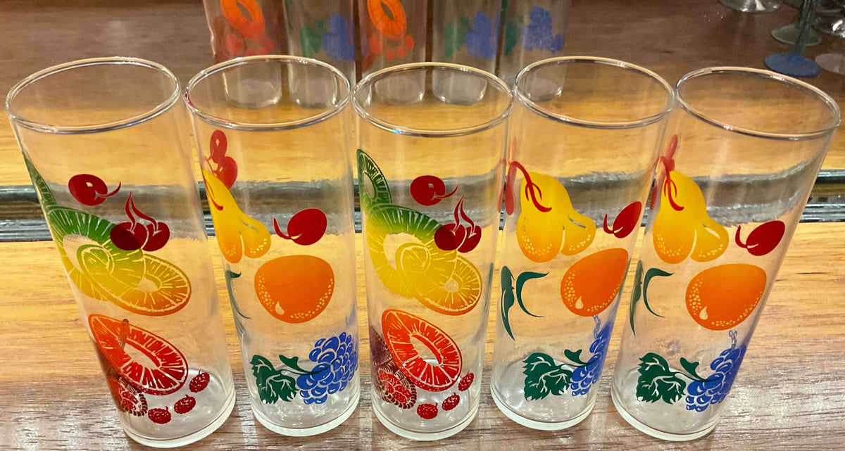 Four clear drinking glasses with colorful fruit prints, including oranges, pears, cherries, and grapes, arranged on a wooden surface.