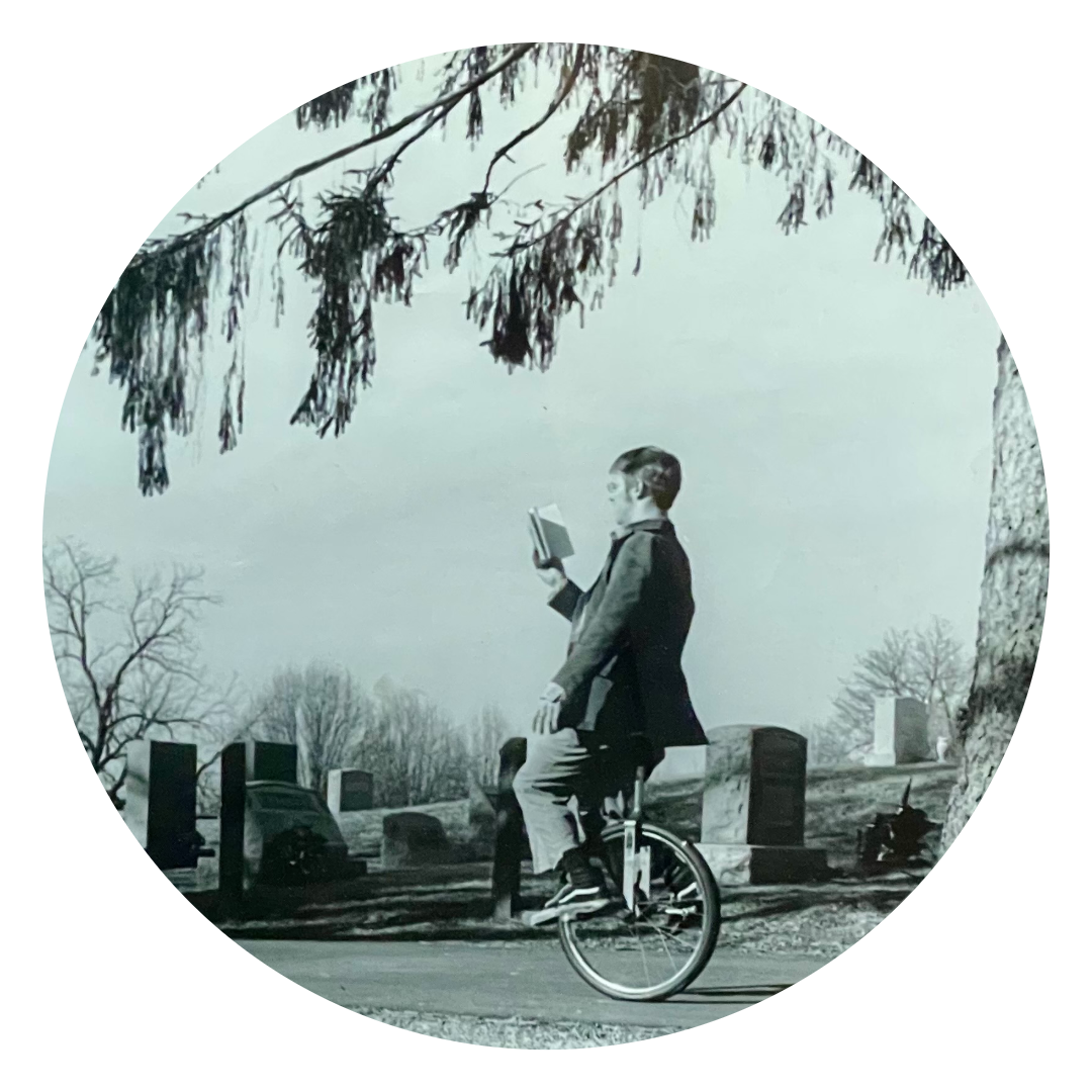 Person on a unicycle reading a book in a graveyard in black and white.