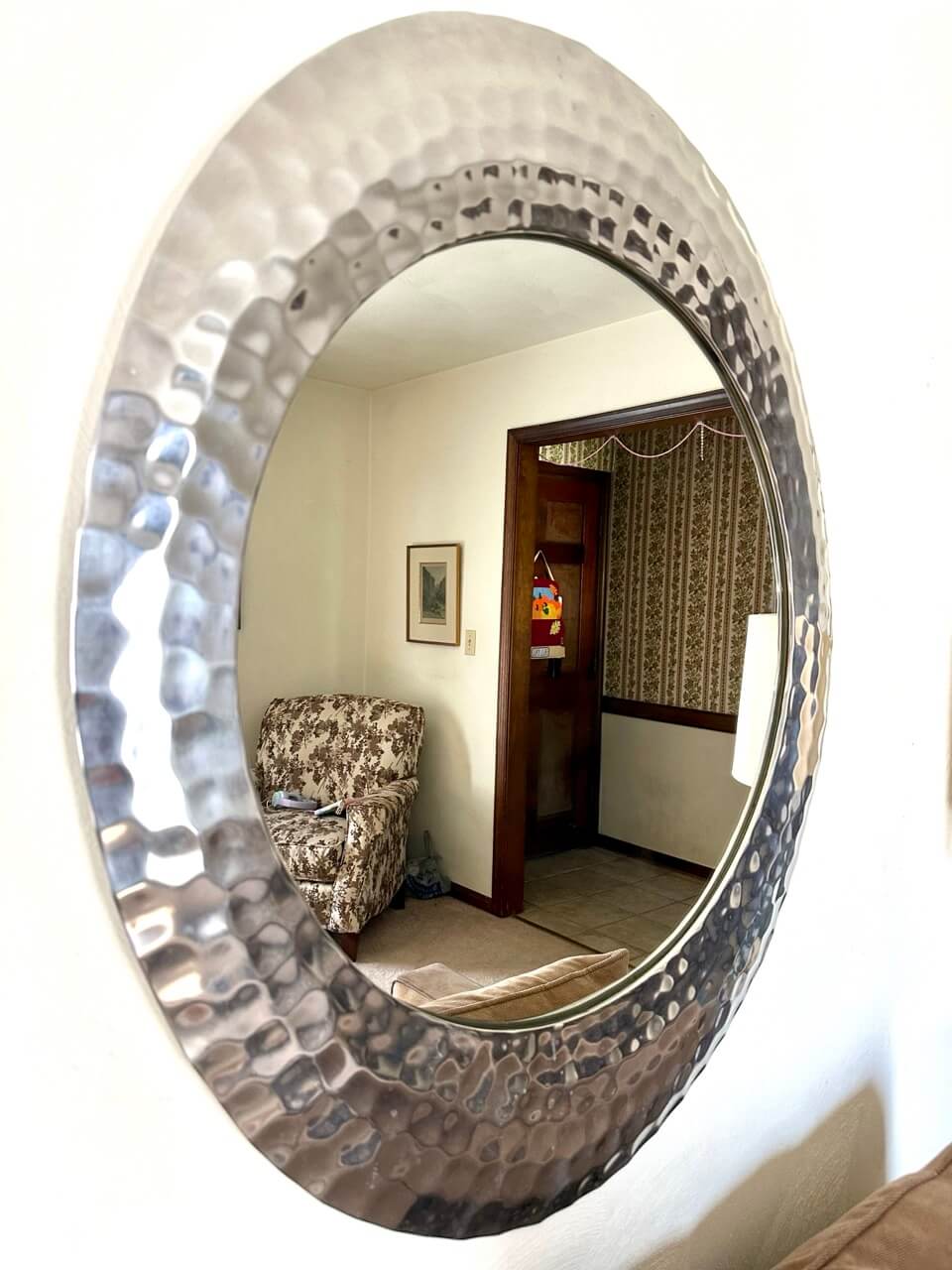 Round metallic mirror reflecting a room with a patterned armchair and door.