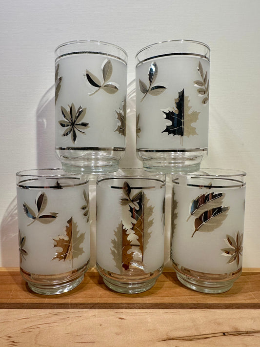 Front view of five frosted clear frosted drinking glasses with etched autumn leaf patterns in metallic silver, arranged in a stacked pyramid display on a wooden surface against a plain light background.
