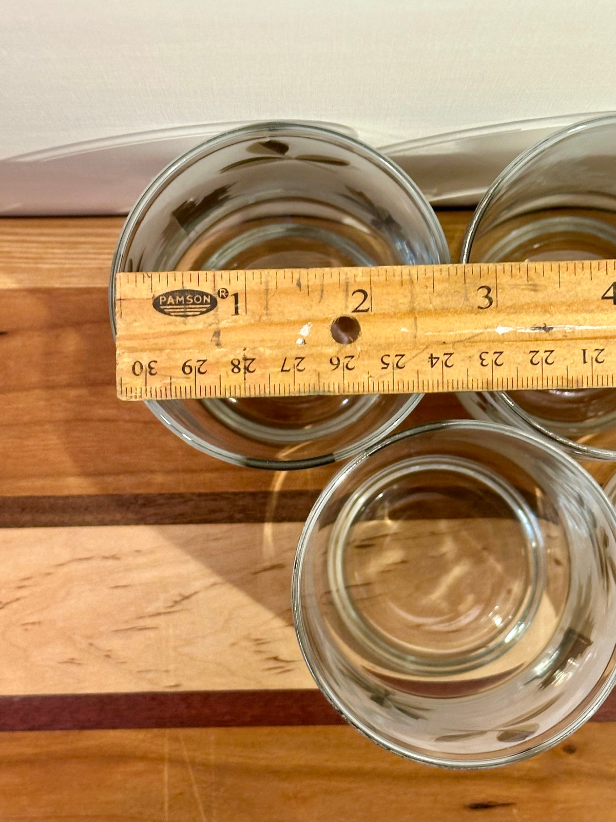 Three clear glass tumblers arranged on a wooden surface with a wooden ruler laid across the top glasses to show their diameter. 