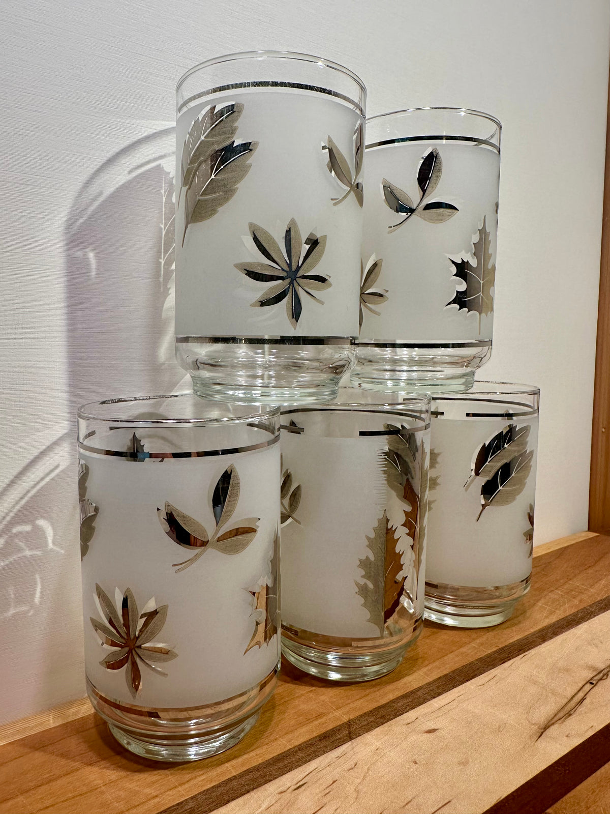 Five frosted glass tumblers side view stacked in pyramid formation with metallic silver botanical leaf patterns, arranged on a wooden shelf against a white wall. 