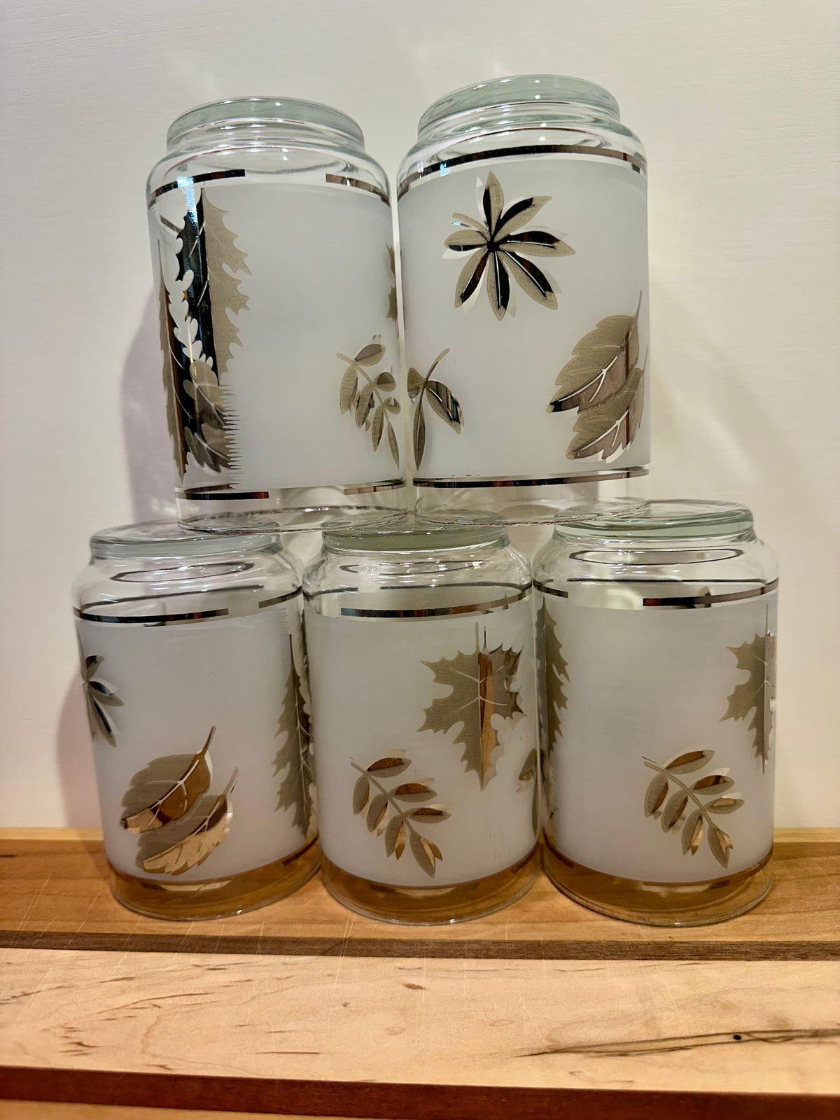 Five frosted glasses arranged in a stacked pyramid display upside down with metallic silver leaf designs, arranged on a wooden surface against a plain light background.