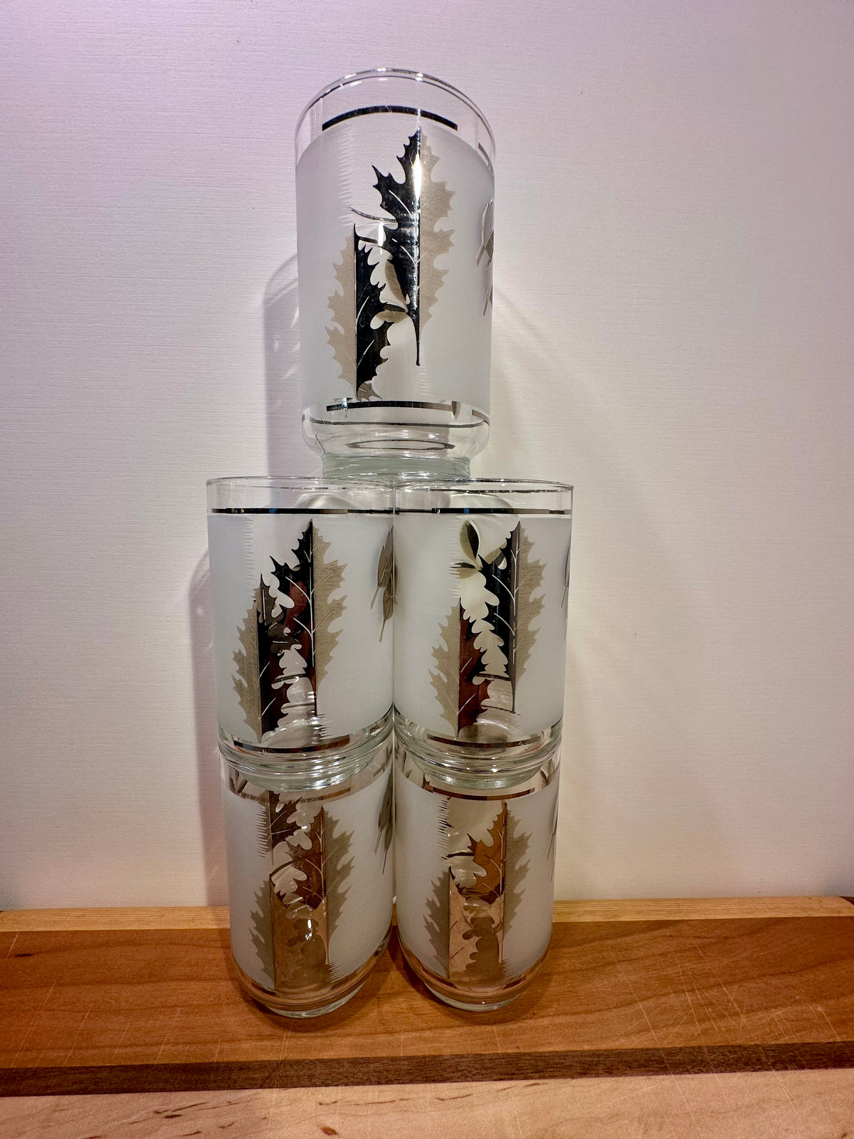 Five clear glass tumblers stacked in a pyramid arrangement on a wooden surface against a plain off-white wall. Each tumbler is decorated with a pressed oak leaf design in silver on frosted clear glass.