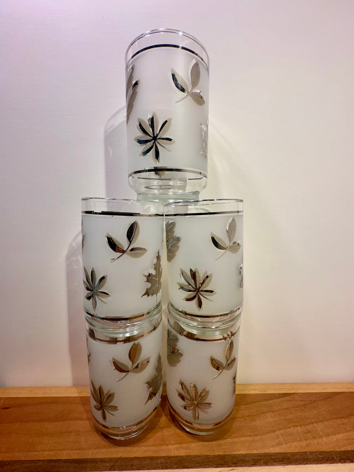Five frosted glass tumblers with metallic silver leaf patterns, stacked in a pyramid arrangement on a wooden surface against a plain white wall.