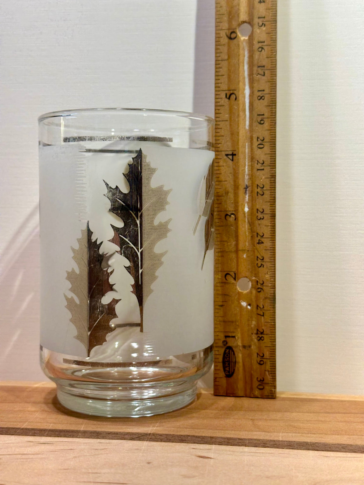 A frosted glass tumbler with a silver oak leaf design with light fading, shown next to a wooden ruler on a light wood surface against a white wall.