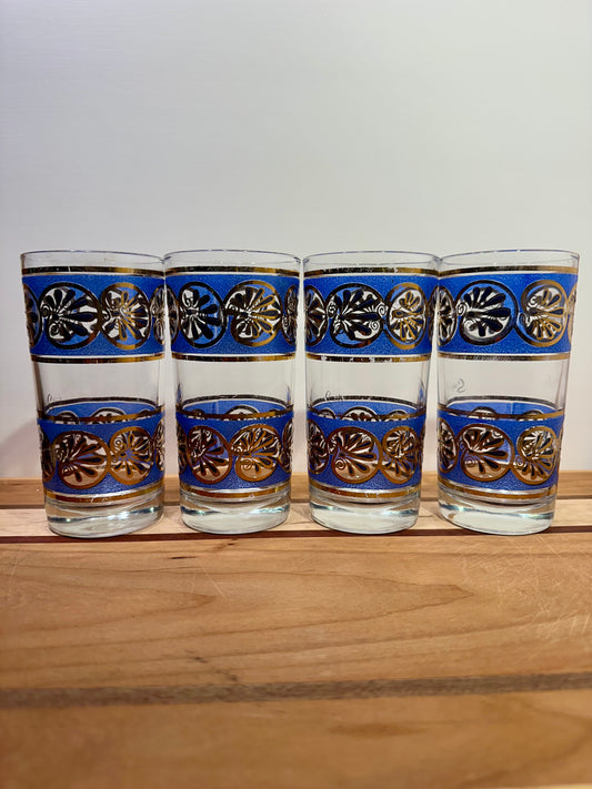 Four clear drinking glasses lined up side by side with blue bands and metallic gold seashell patterns, arranged on a wooden surface against a plain light background.