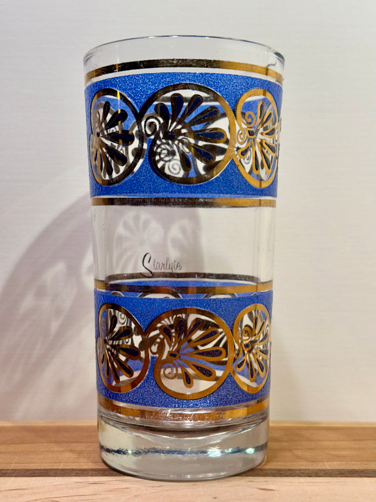 A single clear highball glass tumbler with two decorative bands featuring blue background, gold trim, and seashell motifs, shown on a wooden surface against a light wall.