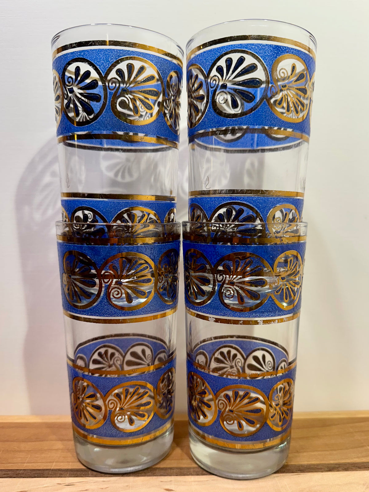 Four tall clear glass tumblers stacked vertically in twos, decorated with blue bands featuring metallic gold seashell scrollwork patterns, set against a plain light background.