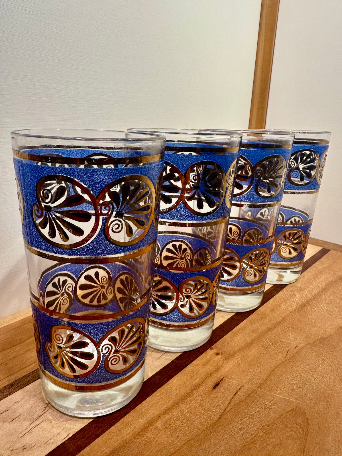 Angled side view of four clear glass tumblers with blue bands and metallic gold and shell patterns, arranged on a wooden surface against a light wall.