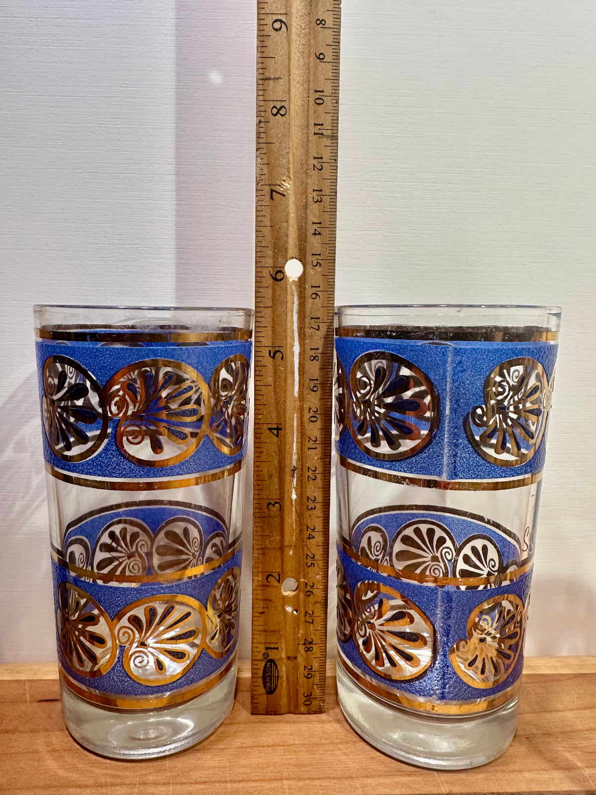 Two clear glass tumblers with blue bands and gold shell cutout patterns, shown on a wooden surface with a wooden ruler standing between them for scale.