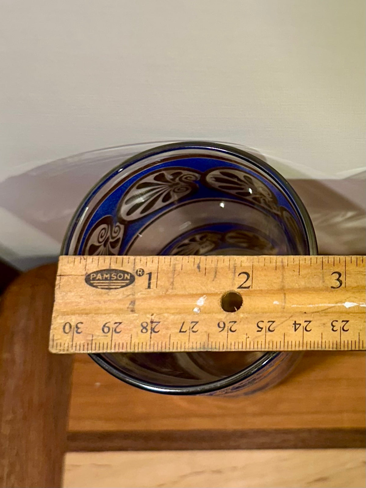 A close-up top view of a blue patterned glass tumbler with a wooden ruler placed across the top to show the diameter, on a wooden surface against a light background.