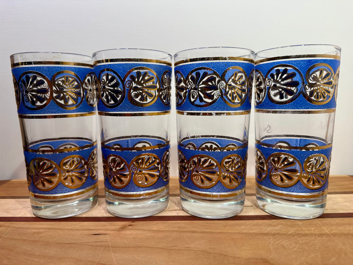 Landscape view of four clear glass tumblers with blue and gold seashell patterns, arranged side-by-side on a wooden surface against a plain light background.