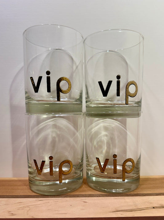 Four clear glass tumblers stacked in a 2x2 grid, each printed with the word 'vip' in gold lowercase letters. The glasses are displayed on a light wooden surface against a plain white wall.