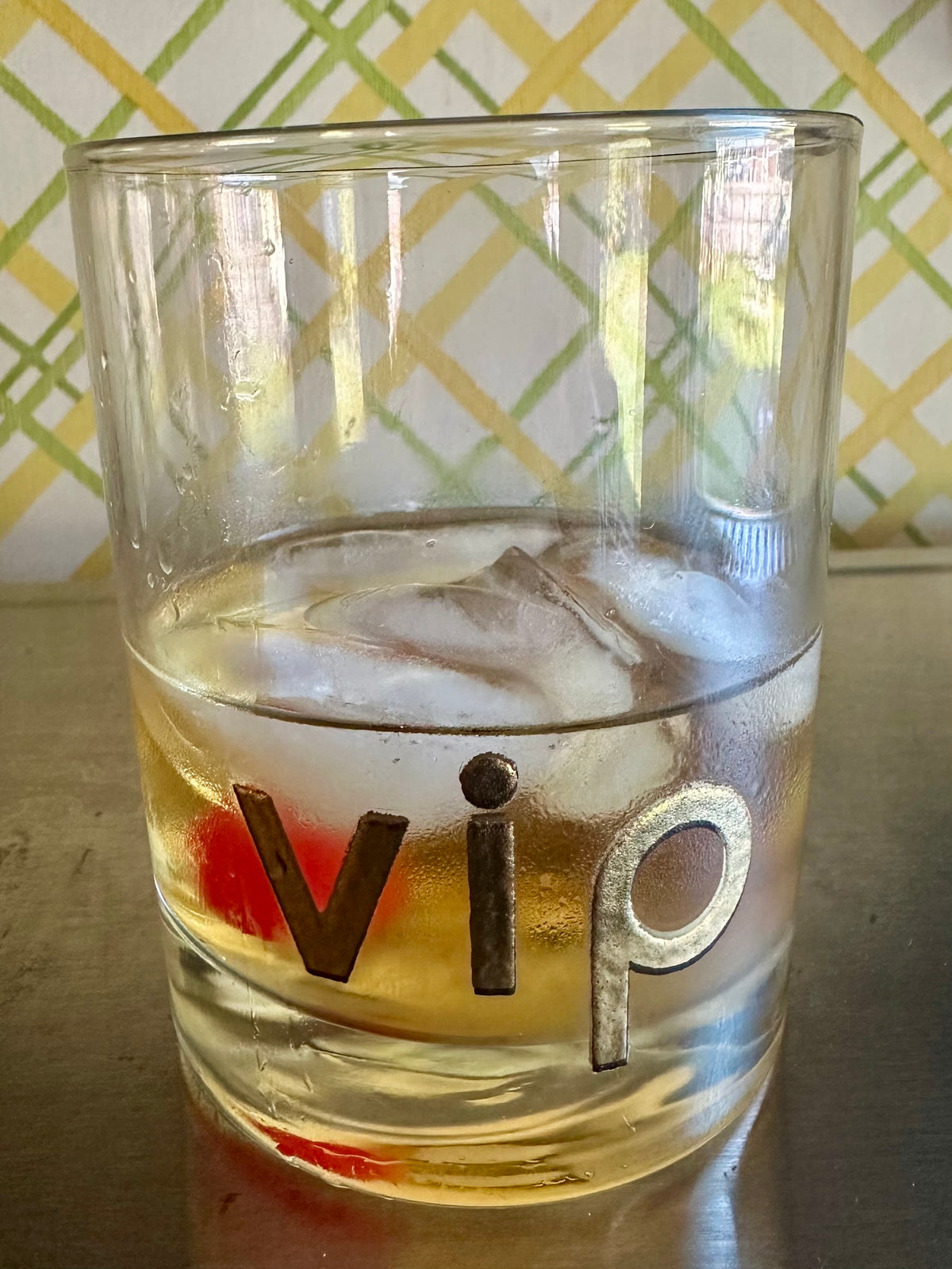 A clear glass rocks tumbler with the letters 'vip' in metallic gold, containing a cold golden drink with ice and a cherry. The glass is set on a reflective metallic surface with a green and yellow patterned background.