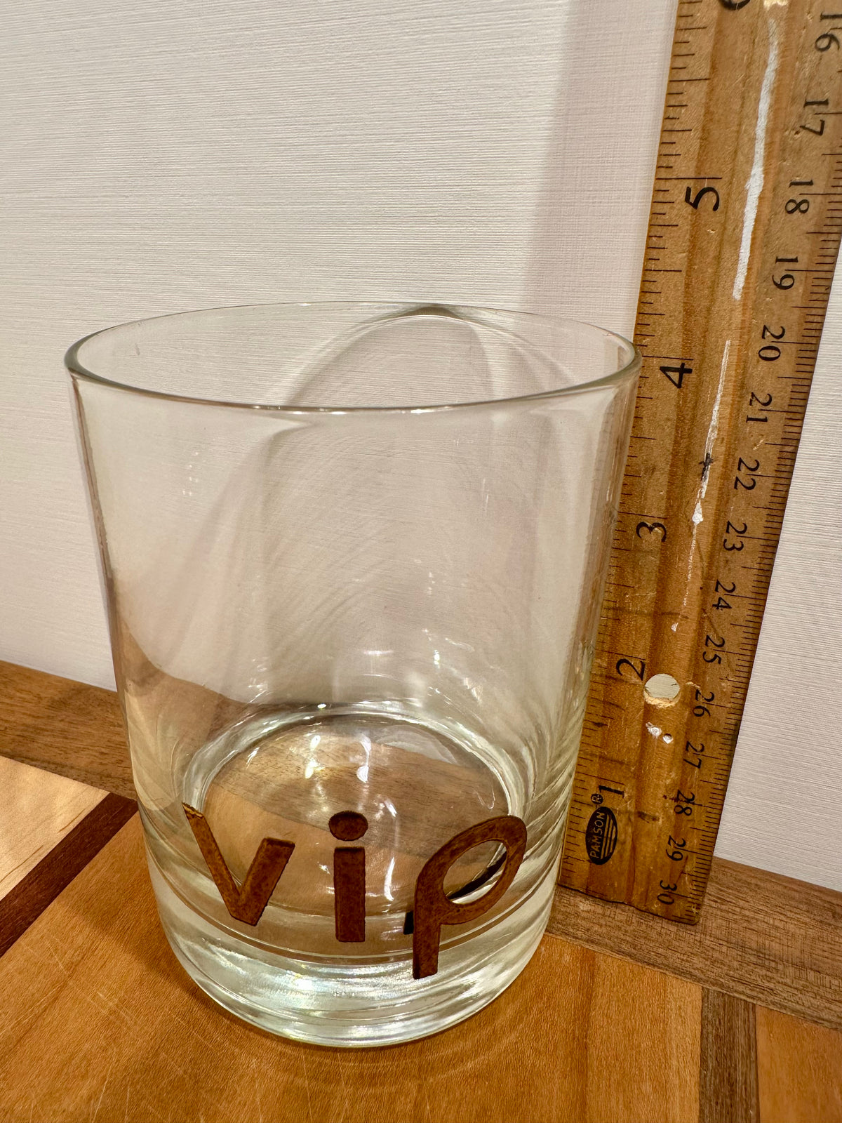 A clear glass tumbler with the letters 'vip' printed in gold on the front, shown on a wooden surface next to a wooden ruler for scale against a white wall.