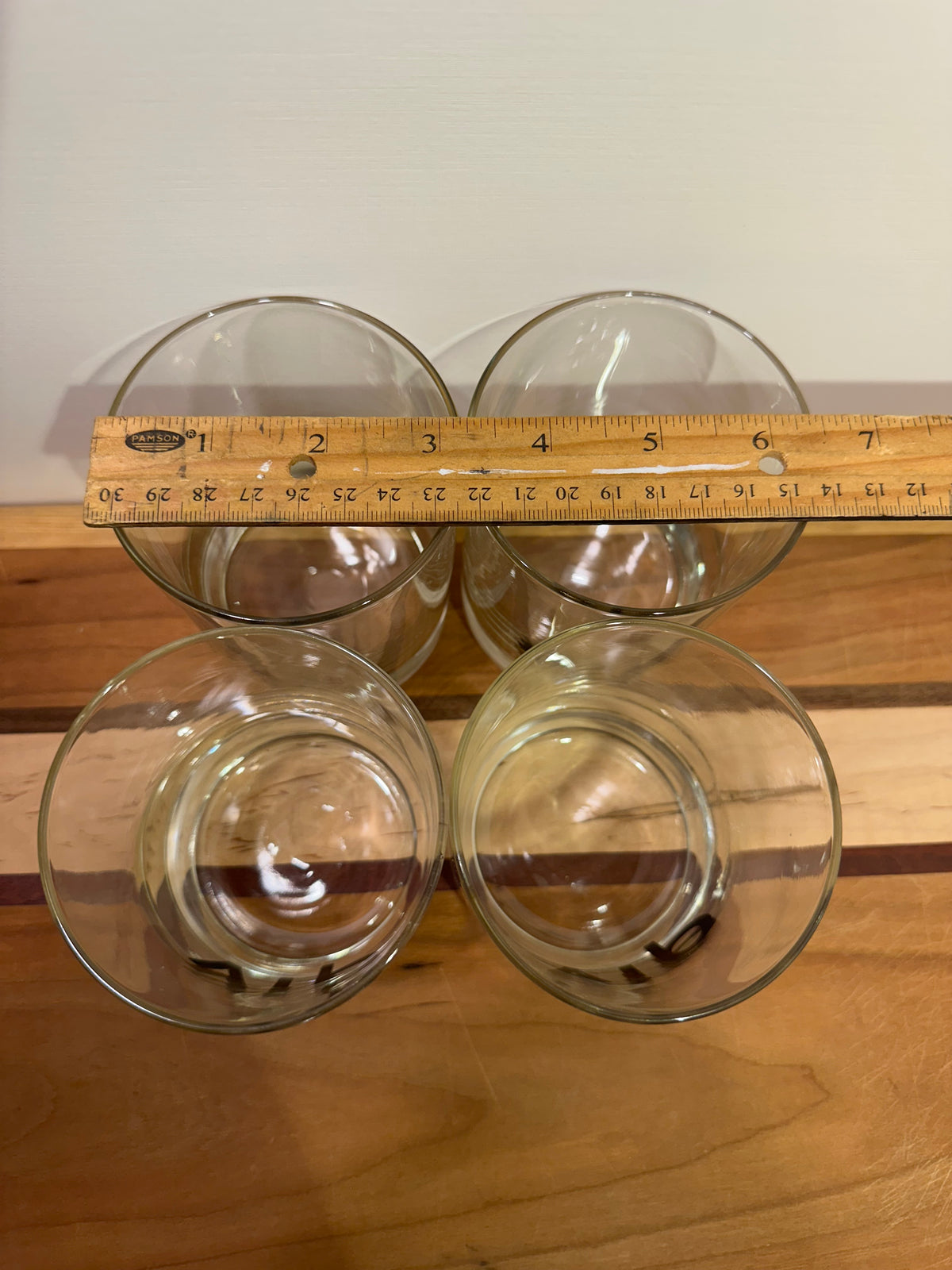 Four clear glass tumblers arranged in a 2x2 grid on a wooden surface in a top down view, with a wooden ruler laid across the top two glasses to indicate rim diameter against a plain light background.
