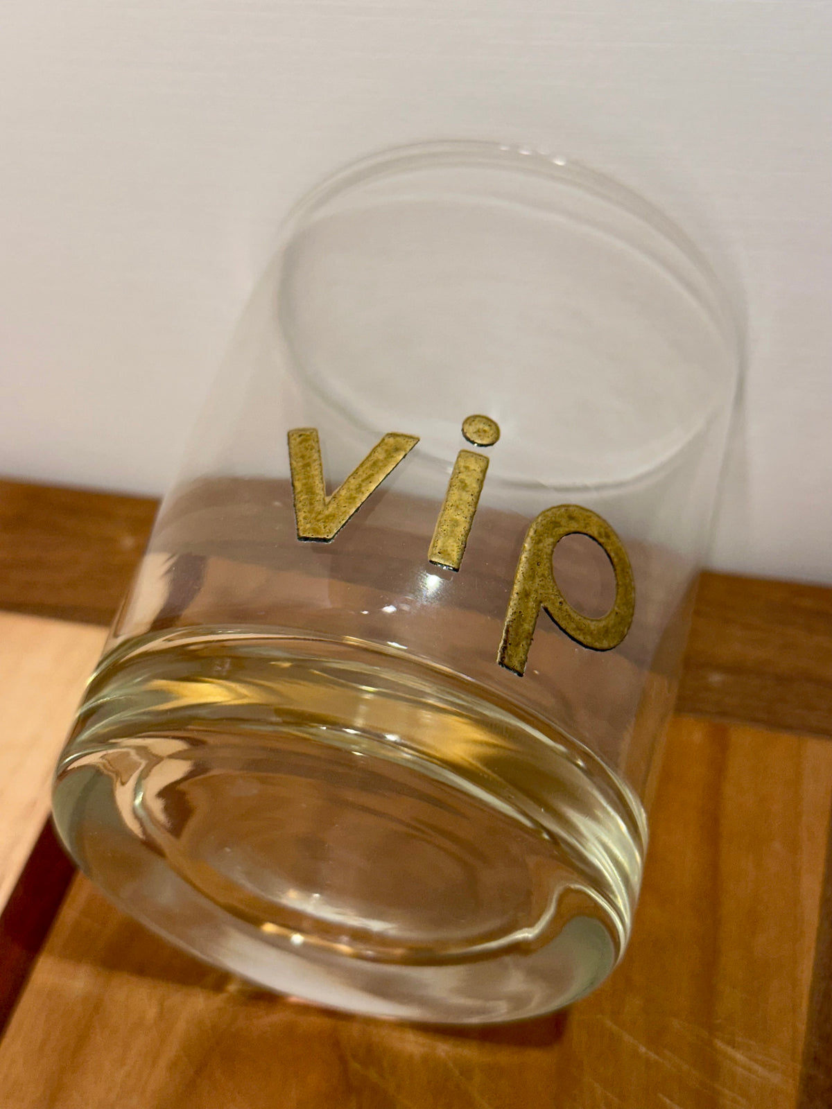 A clear glass drinking tumbler laying on its side with the word 'vip' printed in metallic gold lettering, resting on a wooden surface against a plain light background.