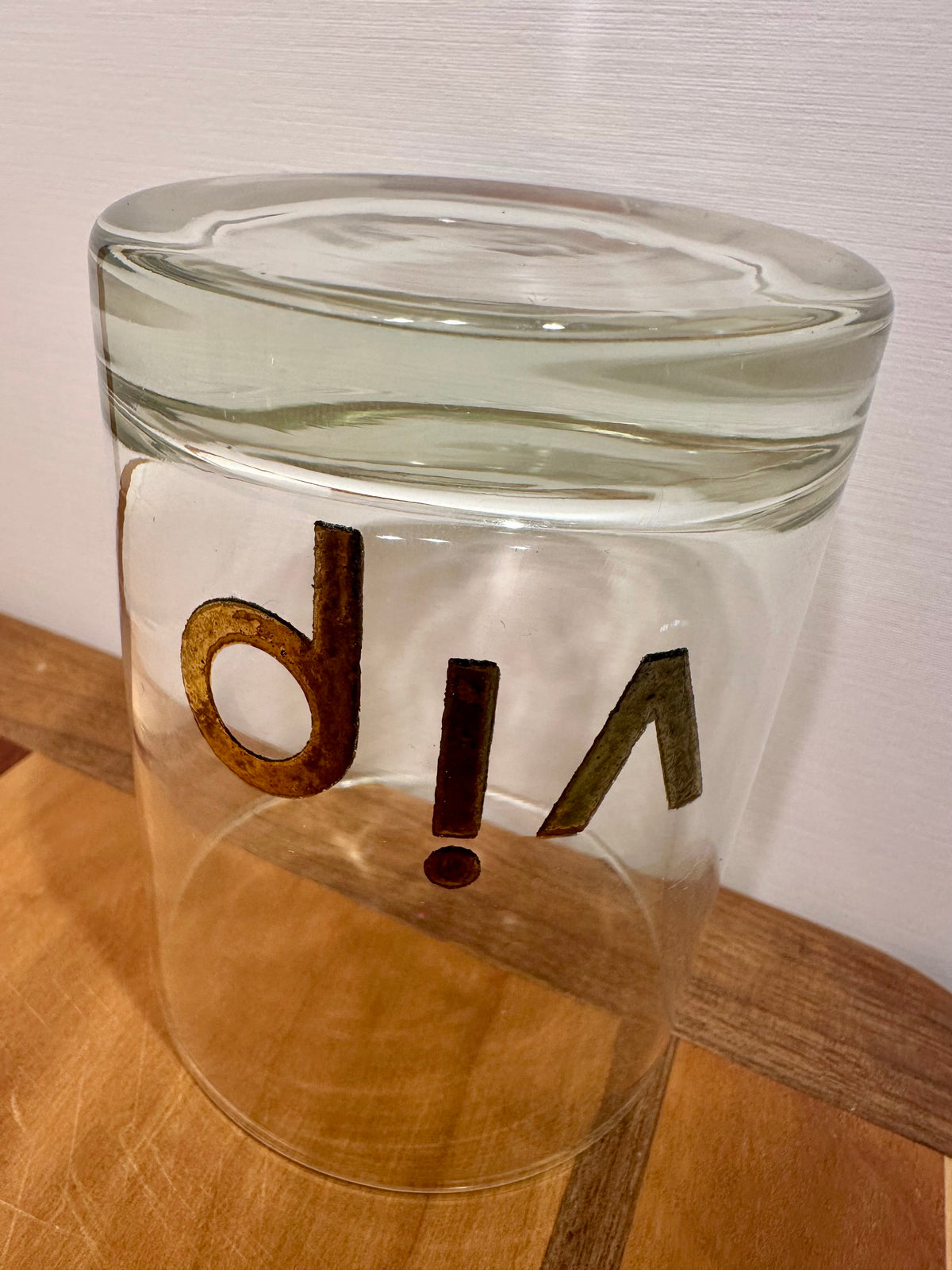 A clear glass tumbler upside down showing a close up of the solid glass base, featuring the letters 'vip' in a gold metallic font on the front, resting on a wooden surface against a light wall.