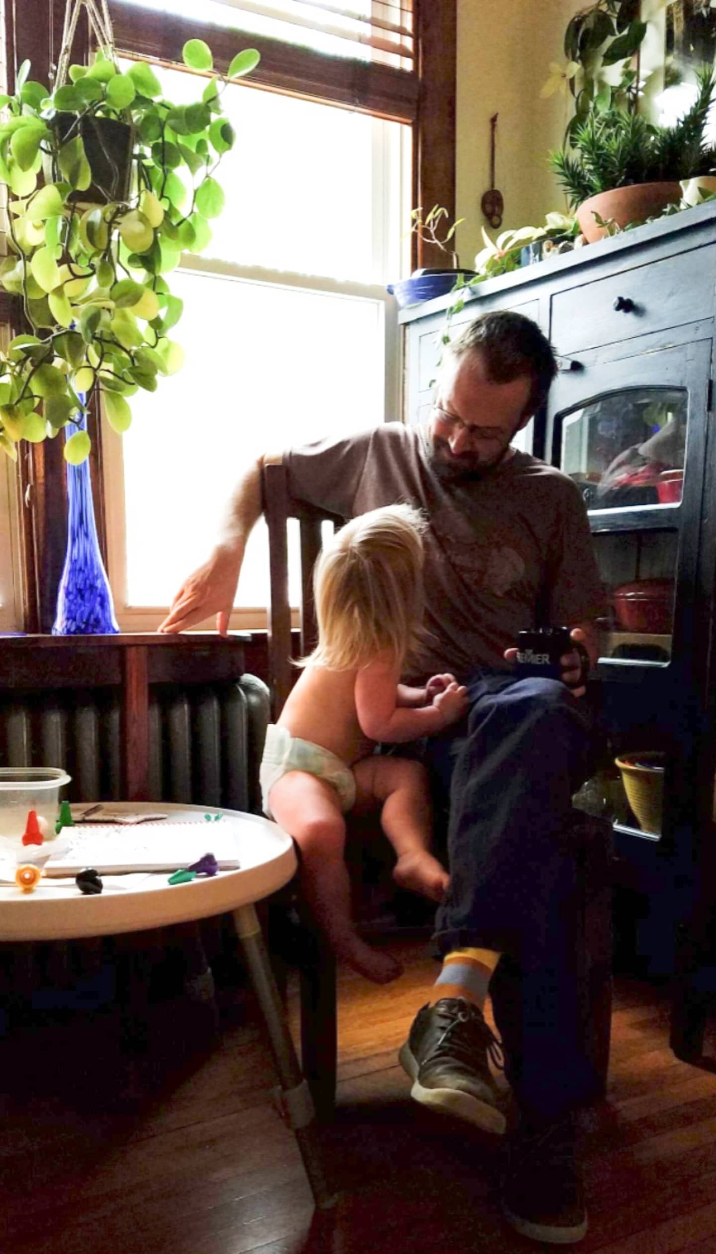 Man sitting with a child on a chair in a room with plants and a window.